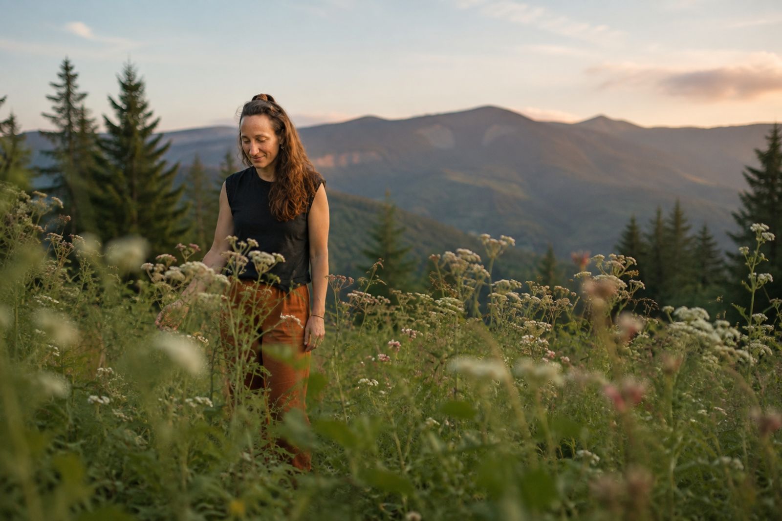 Eine Frau geht bei Sonnenuntergang durch eine Bergwiese mit weißen und rosa Wildblumen. Im Hintergrund Nadelbäume und Bergketten im Abendlicht.