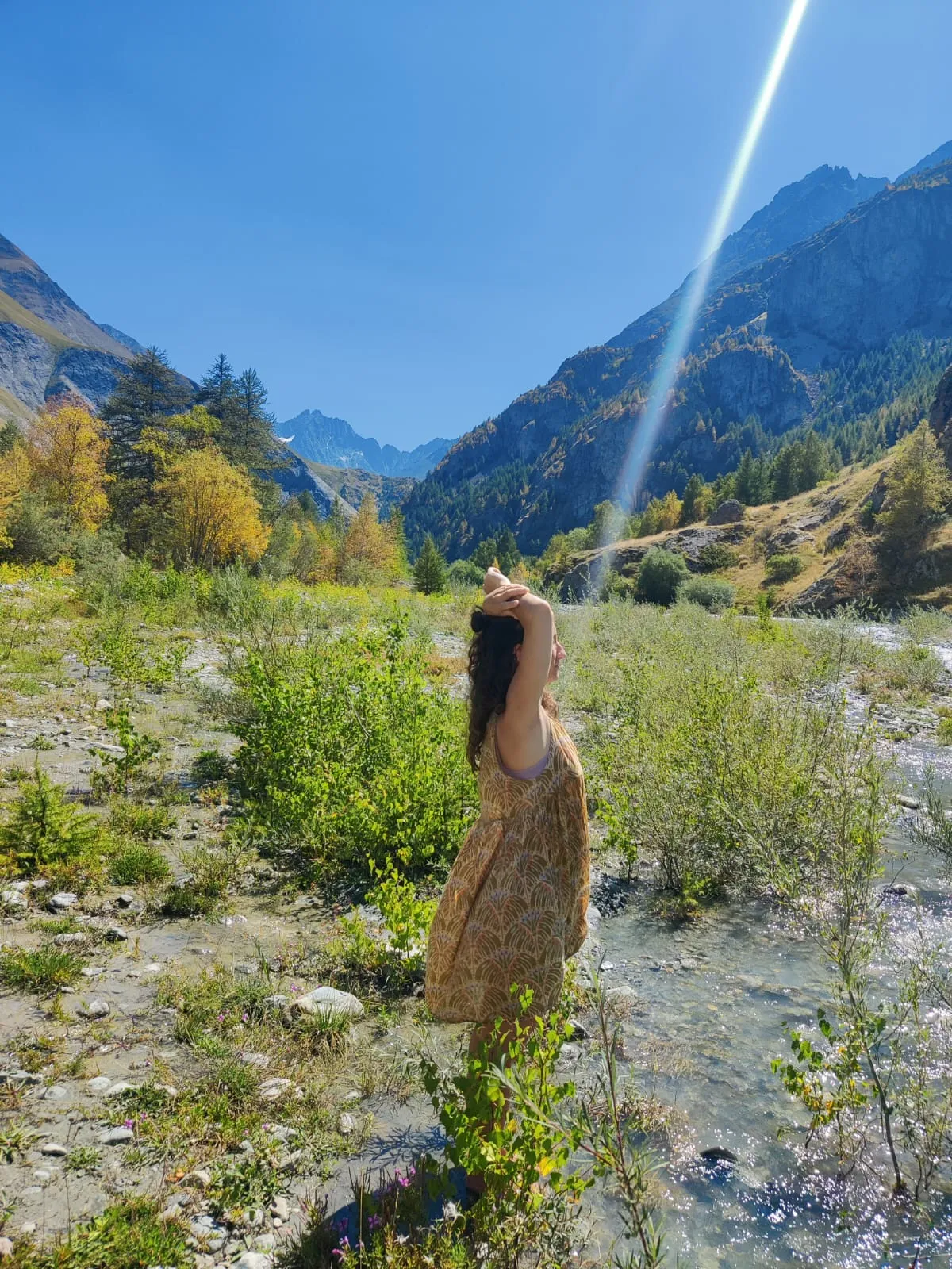 Eine Frau steht in einem Bachbett in einem Bergtal, umgeben von Sträuchern, dahinter Felswände und herbstlich gefärbte Lärchen.