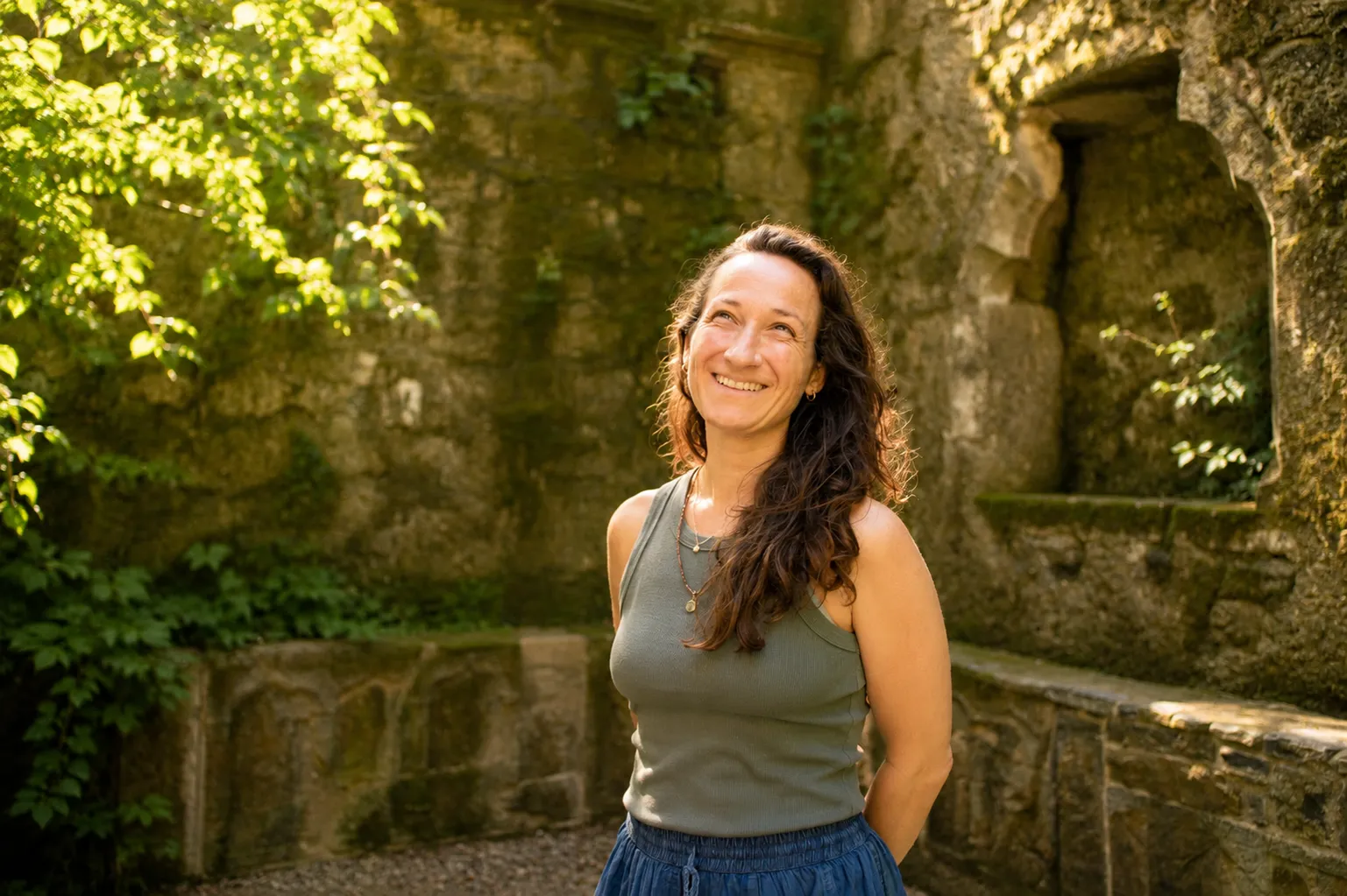 Valerie lacht und blickt nach oben, vor einer bemoosten alten Steinmauer.