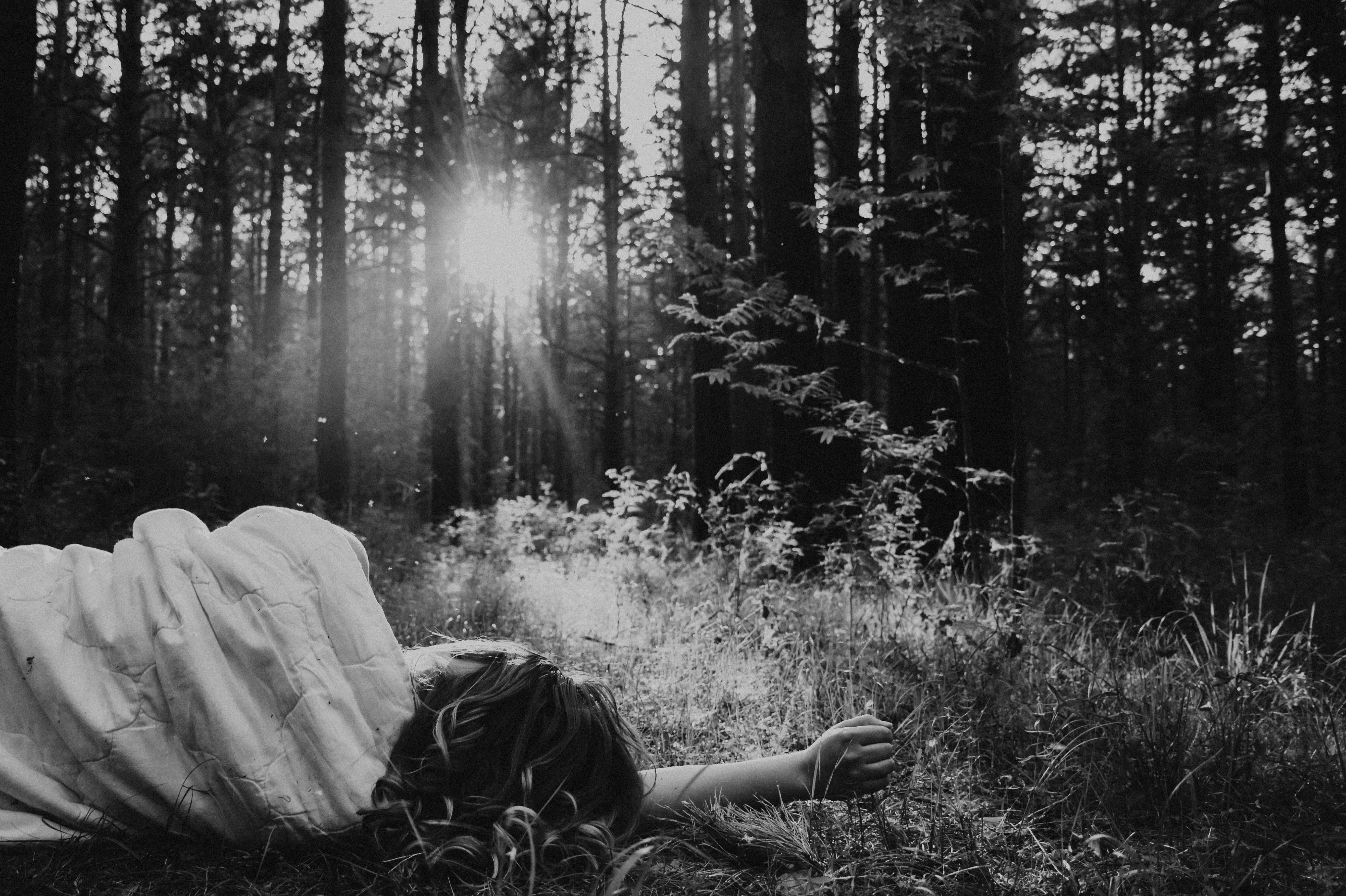 Eine Frau liegt unter einer hellen Decke im Gras eines Nadelwaldes, die Sonne steht tief zwischen den Stämmen. Schwarzweiß.