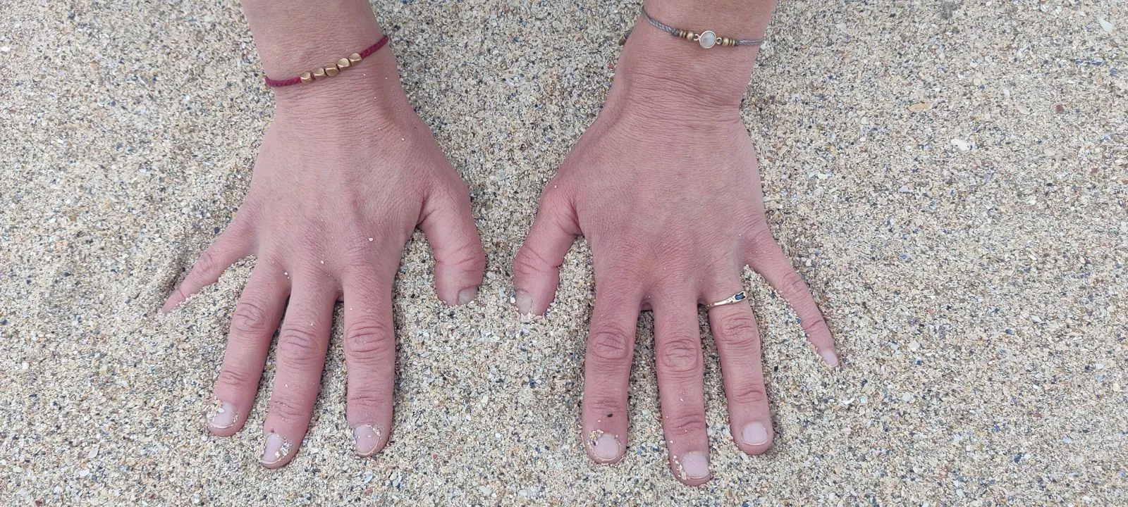 Zwei Hände liegen mit gespreizten Fingern flach im hellen Sand, an jedem Handgelenk ein schmales Armband, an einem Finger ein Ring.